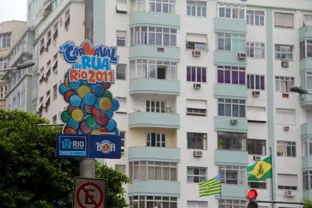 A vibrant street sign for Carnaval de Rua Rio 2011 with colorful balloons stands in front of a high-rise building adorned with green and white flags. At this bustling intersection, traffic lights guide the flow of festival-goers immersed in the Carnaval spirit.