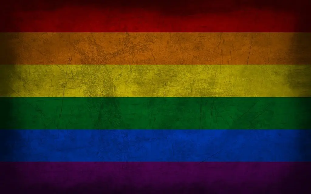 A grunge-style rainbow flag with horizontal stripes in red, orange, yellow, green, blue, and purple. The texture gives the appearance of worn, faded fabric.