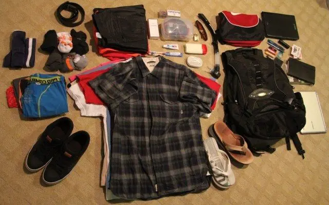 A variety of travel items are neatly arranged on a beige carpet, showcasing impeccable packing light techniques. The collection includes shirts, shorts, socks, and essentials like flip-flops, a backpack, and toiletries—all you need for the perfect trip without ever having to check a bag.