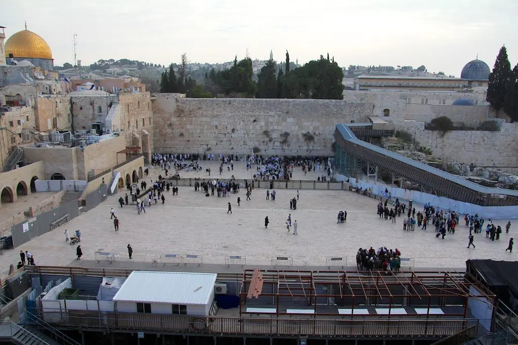 A distant view of the Western Wall plaza in Jerusalem, a prelude to the sacred gatherings in the Holy Land. Groups of people are gathered near the wall, while the Dome of the Rocks golden dome gleams subtly in the overcast sky.