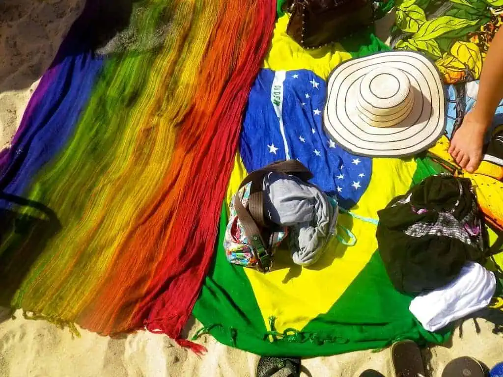 Colorful beach towels and a vibrant Brazilian flag towel, alongside a traditional canga, are laid out on the sand. A wide-brimmed hat, bags, and clothing add to the scenes charm. Sandals peek out at the bottom, completing this picturesque beach setup.
