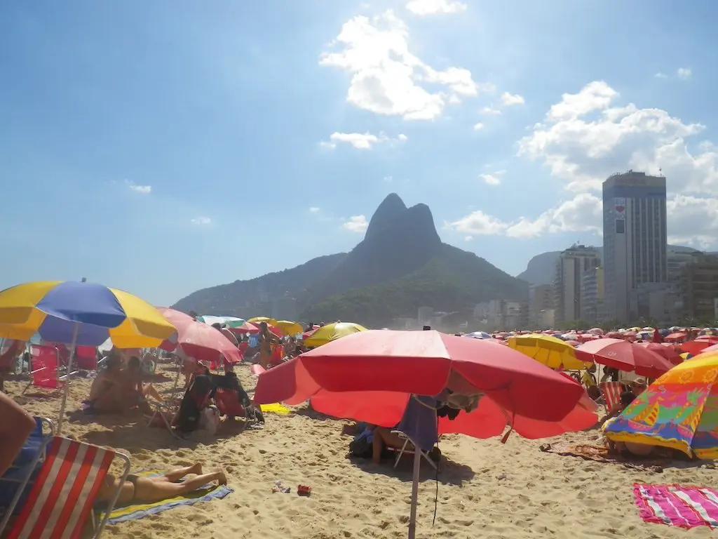 A sunny Brazilian beach scene with colorful umbrellas and sunbathers practicing perfect etiquette on the sand. In the background, a mountain range with a distinctive peak and city buildings are visible against a blue sky with scattered clouds.