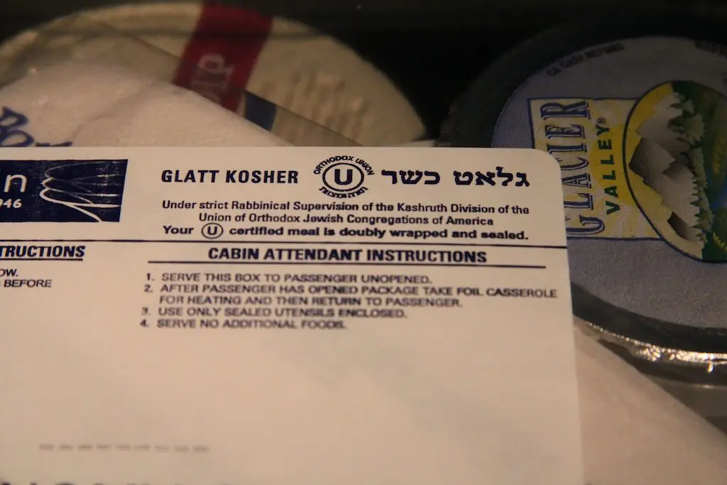 A close-up of a paper label titled Glatt Kosher highlights kosher food certification by the Orthodox Union, featuring meal heating instructions. The kosher symbol and partially visible packaged food items complete the display.
