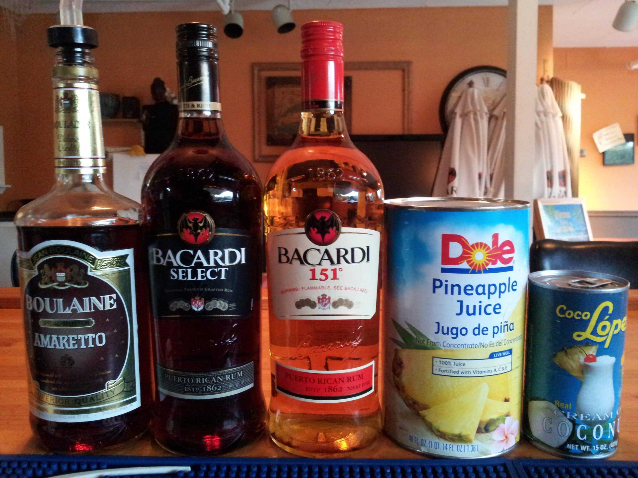 Bottles of Boulaine Amaretto, Bacardi Select, and Bacardi 151 rum—dubbed the perfect ingredients for rocket fuel cocktails—are lined up next to a can of Dole pineapple juice and Coco López cream of coconut on a wooden countertop. Various items are blurred in the background.