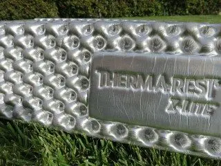 Close-up of a silver, textured foam sleeping pad with Therm-a-Rest Z Lite Sol embossed on it. Resting on green grass, the pad shines subtly against a blurred backdrop of lush bushes.