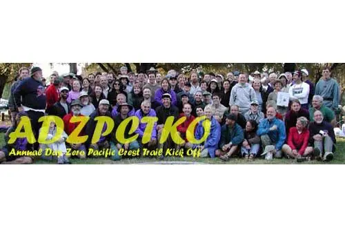 A large group gathers for a photo at the Annual Day Zero Pacific Crest Trail Kick Off event, ADZPCTKO emblazoned in vibrant text across the scene. Towering trees create a picturesque backdrop.