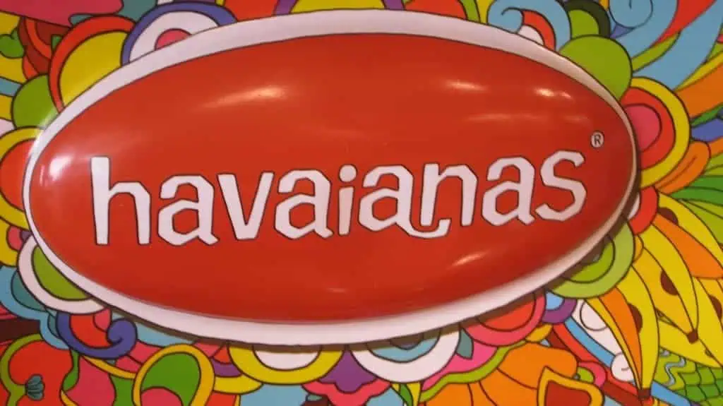A large red oval sign prominently displays the word havaianas in striking white lowercase letters. The vibrant background bursts with colorful and abstract patterns, featuring swirling floral elements in bright hues of yellow, green, orange, pink, and blue.