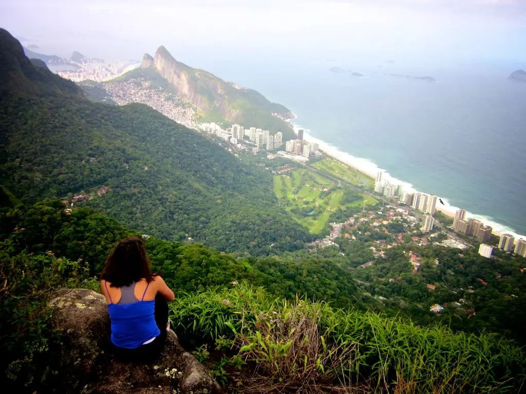 A person with long hair and a blue top sits on a rocky ledge, embodying the spirit of hikes in Rio de Janeiro. They overlook a lush green valley, with the sprawling city, majestic mountains, and vast ocean in the distance under a cloudy sky.
