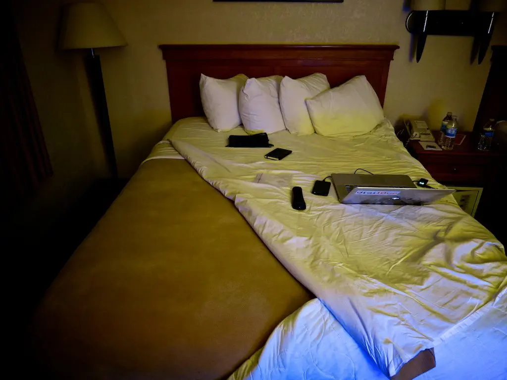 A dimly lit hotel room captures the essence of Night Before the Morning. The unmade bed, with four pillows and a light brown blanket pulled back, holds a projector, remote controls, and a phone. Beside it, a lamp casts shadows over a nightstand cluttered with various items.