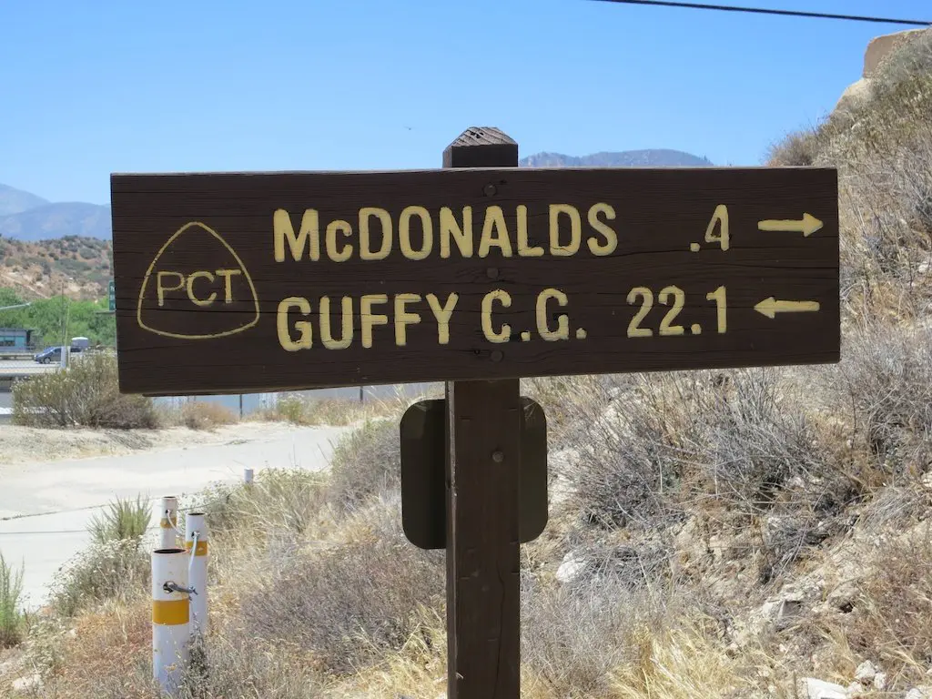 In the warm afternoon sun, a wooden trail sign with the PCT symbol stands amid an arid landscape. Arrows guide hikers to McDonalds 0.4 miles ahead and Guffy C.G. 22.1 miles away, with rugged mountains painting the horizon.