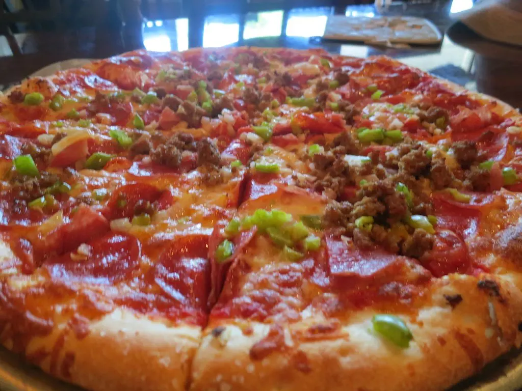 A delicious pizza topped with pepperoni, sausage, green bell peppers, and cheese sits enticingly on a metal tray. Sliced to perfection, it beckons from the foreground of a bustling restaurant scene—a zero-second pause before you dive in.