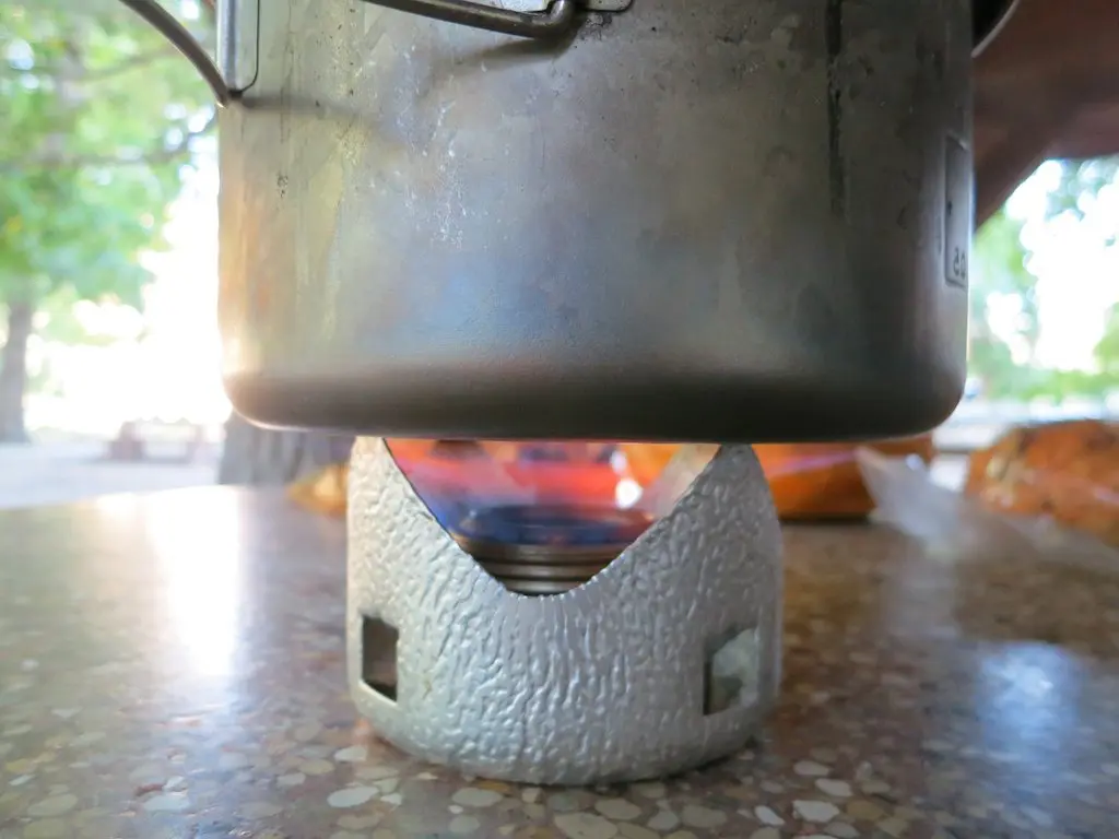 A metal pot sits on a small camping stove, its flame flickering against a speckled outdoor surface. Blurred trees suggest this scene of culinary adventure unfolds amid the rugged beauty encountered while cooking on the Pacific Crest Trail.