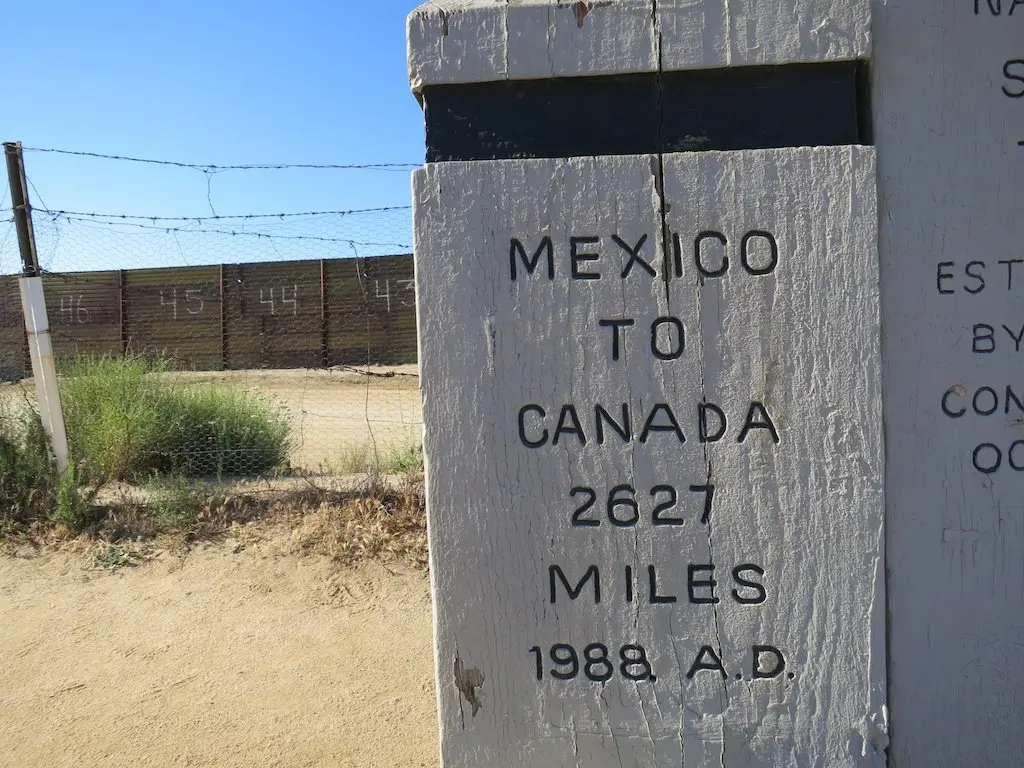 A weathered sign marks the start of PCT Day 1, stretching from Mexico to Canada, indicating 2,627 miles and dated 1988 A.D. It stands near a barbed wire fence and a dirt path under a clear blue sky.