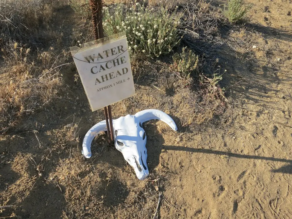 A sign on the dirt path reads Water Cache Ahead, Approx 1 Mile, a welcome message for weary PCT checkers. Beside it lies a white animal skull with horns, embedded in the ground amidst sparse vegetation.