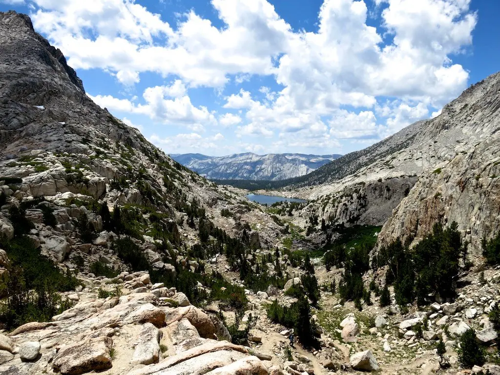 A rugged mountain landscape with rocky terrain and sparse vegetation unfolds as a sneak peek into natures grandeur. A small lake in the Sierra glimmers in the distance, surrounded by hills. Fluffy white clouds dot the bright blue sky, completing this serene vista.