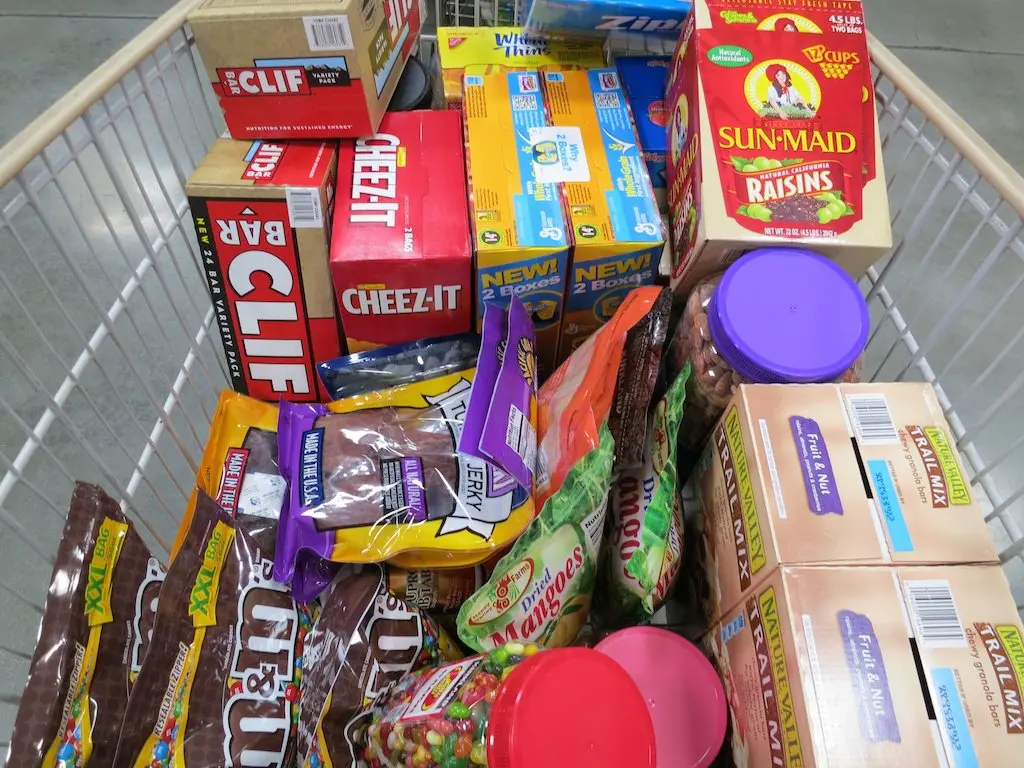 A shopping cart filled with various snacks and groceries, including Clif Bars, Cheez-It crackers, Sun-Maid raisins, beef jerky, M&Ms, and boxed juice pouches. The neatly stacked items provide the perfect resupply for your pantry needs.