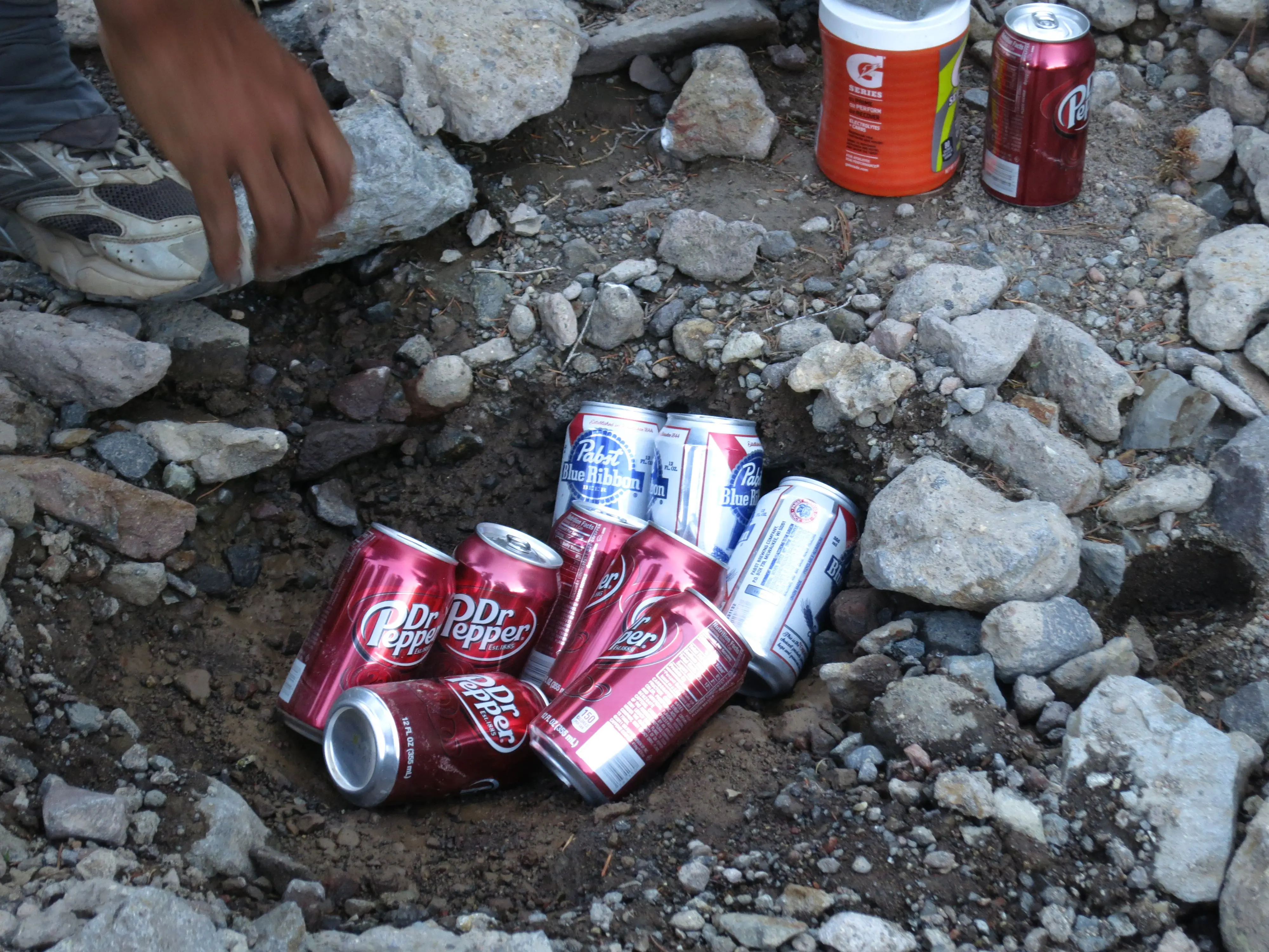 Several soda cans, including Dr Pepper and Pabst Blue Ribbon, rest in a depression surrounded by rocks. A Thru-Hikers hand hovers nearby, with a glimpse of their sneaker on the left. In the background, a Gatorade bottle hints at the refreshment sought by weary travelers of the PCT.