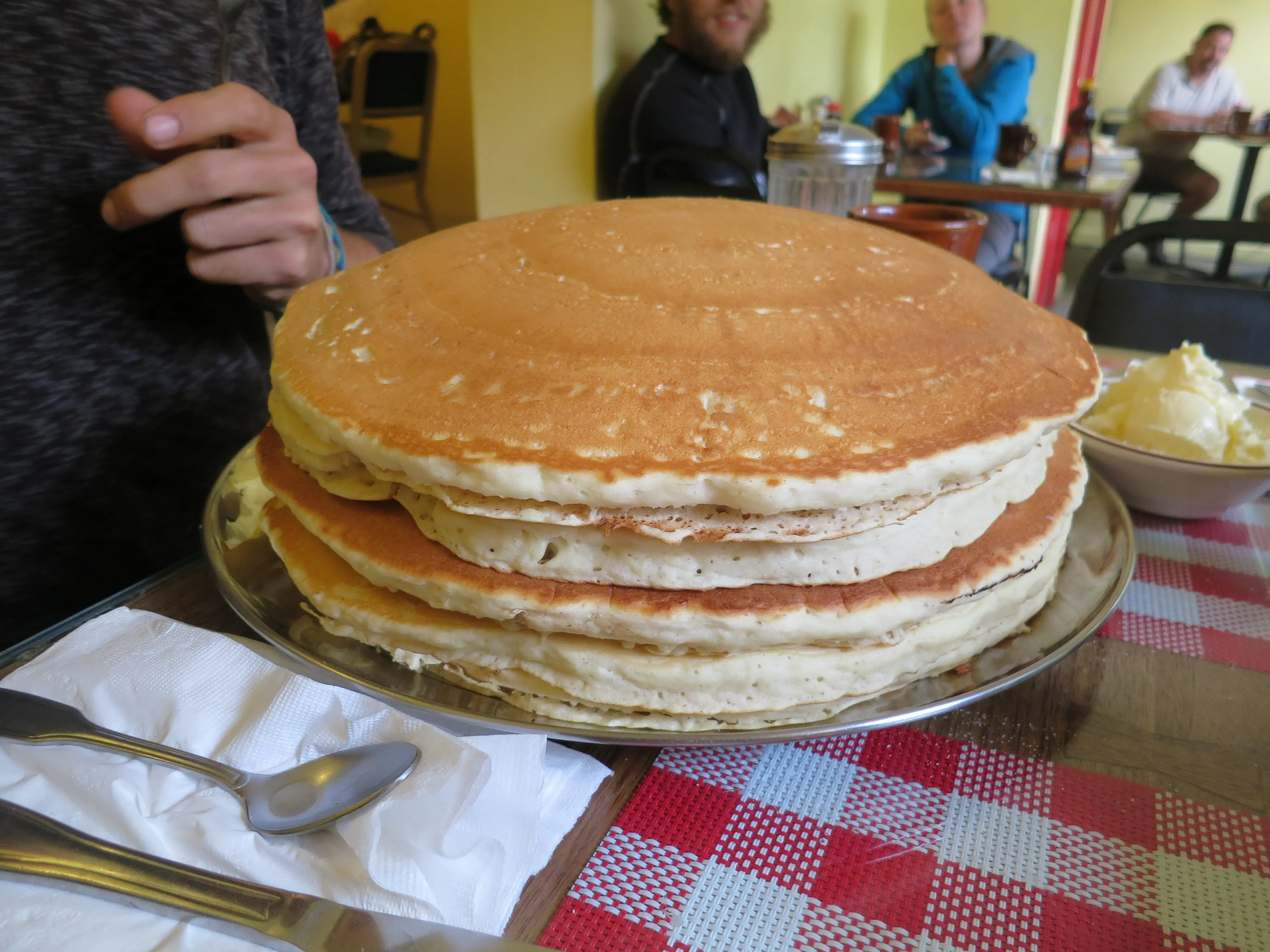 A towering stack of oversized pancakes on a metal plate sits on a red and white checkered tablecloth, reminiscent of a hikers hearty meal after a day on the trails. Utensils and a napkin are beside it, while people gather at tables in the cafes lively background.