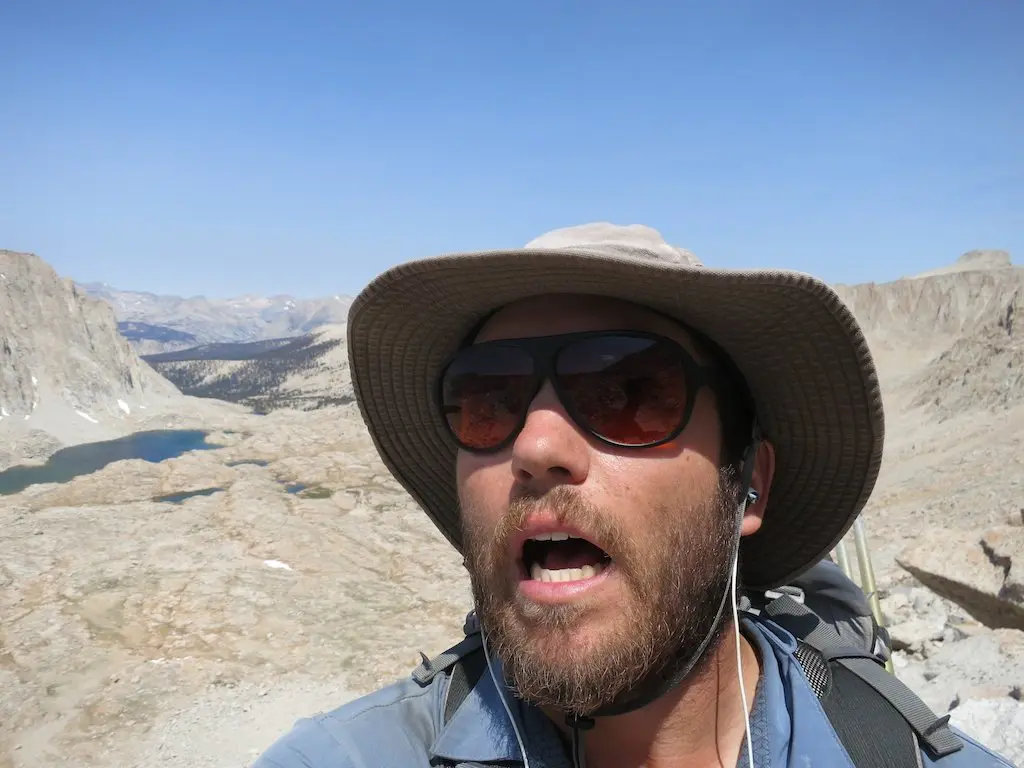 Donning a wide-brimmed hat, sunglasses, and earphones, a hiker snaps a selfie along the rugged PCT. With an open mouth as if singing to the mountains and small lakes below, theyre intent on keeping busy while immersing themselves in natures solitude and beauty.