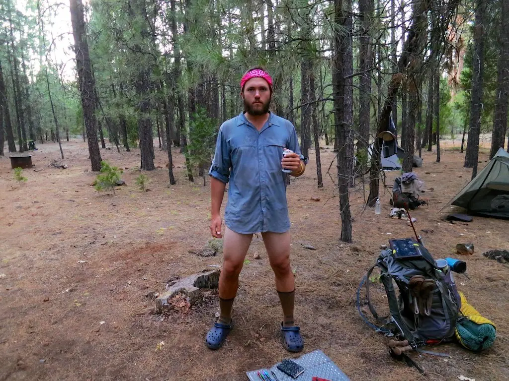 A man stands in a forest clearing, wearing a blue shirt and red headband, with socks and Crocs. He holds a cup amid camping gear like a backpack and tent. The trees hint he might be 600 miles into his PCT journey, ready for some small talk about the trail.