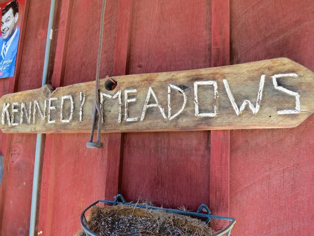 A wooden sign with KENNEDY MEADOWS carved into it is mounted on a red wall. A large nail pierces one of the letters, while below hangs a metal basket brimming with dried plant material. To the left, a partial poster adds to the rustic charm of this Kennedy Meadows tribute.