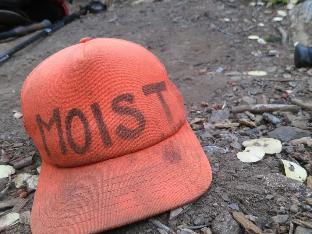 An orange cap with the word MOIST written on it lies on the ground surrounded by dirt, small twigs, and leaves, as if marking a hidden spot along the Pacific Crest Trail.