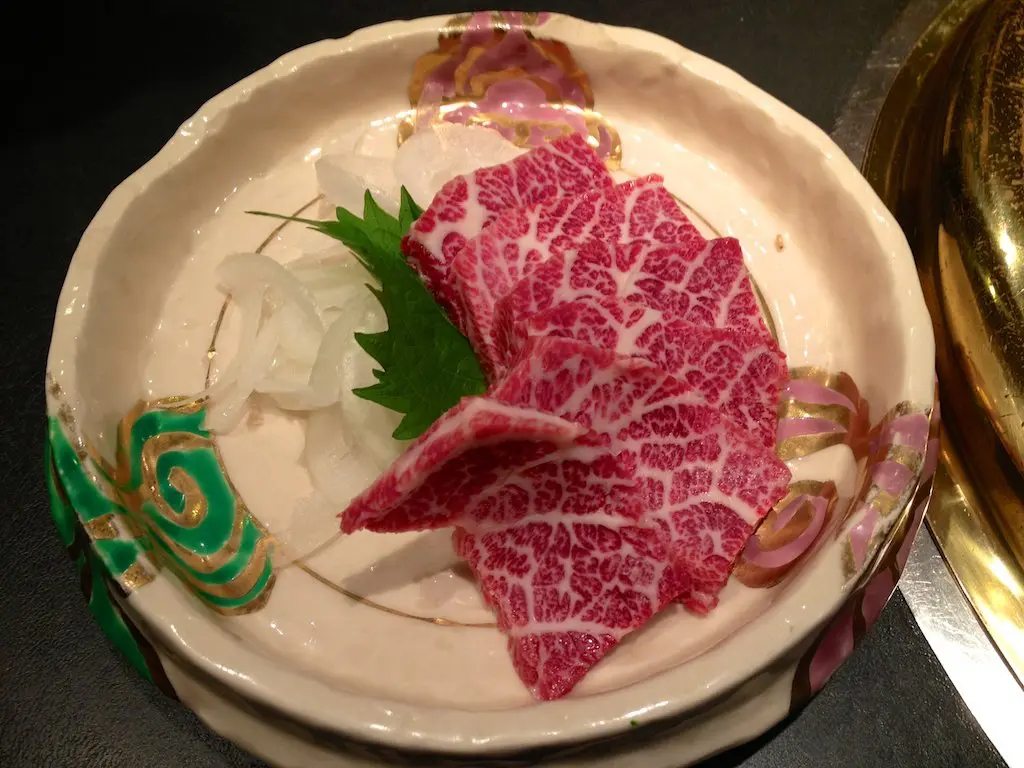 A plate of marbled basashi, featuring raw beef slices garnished with a green shiso leaf and thinly sliced white onions, served on a decorative ceramic dish.