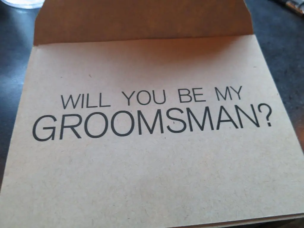A greeting card with the text Will you be my groomsman? in bold, black lettering on a plain, brown background invites an answer to what some consider absurd questions. The card is slightly open, resting on a dark surface.