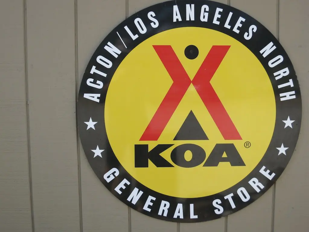 A circular sign with a yellow background features the KOA logo in red and black. The surrounding black border reads ACTON/LOS ANGELES NORTH GENERAL STORE in white letters, reminding campers that sometimes, bigger isnt better when it comes to finding the perfect spot.