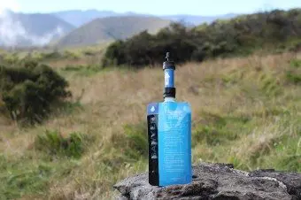 A blue water filtration pouch with a black Sawyer Mini filter attachment rests on a rock in a grassy landscape. Mountains and cloudy skies provide a stunning backdrop.