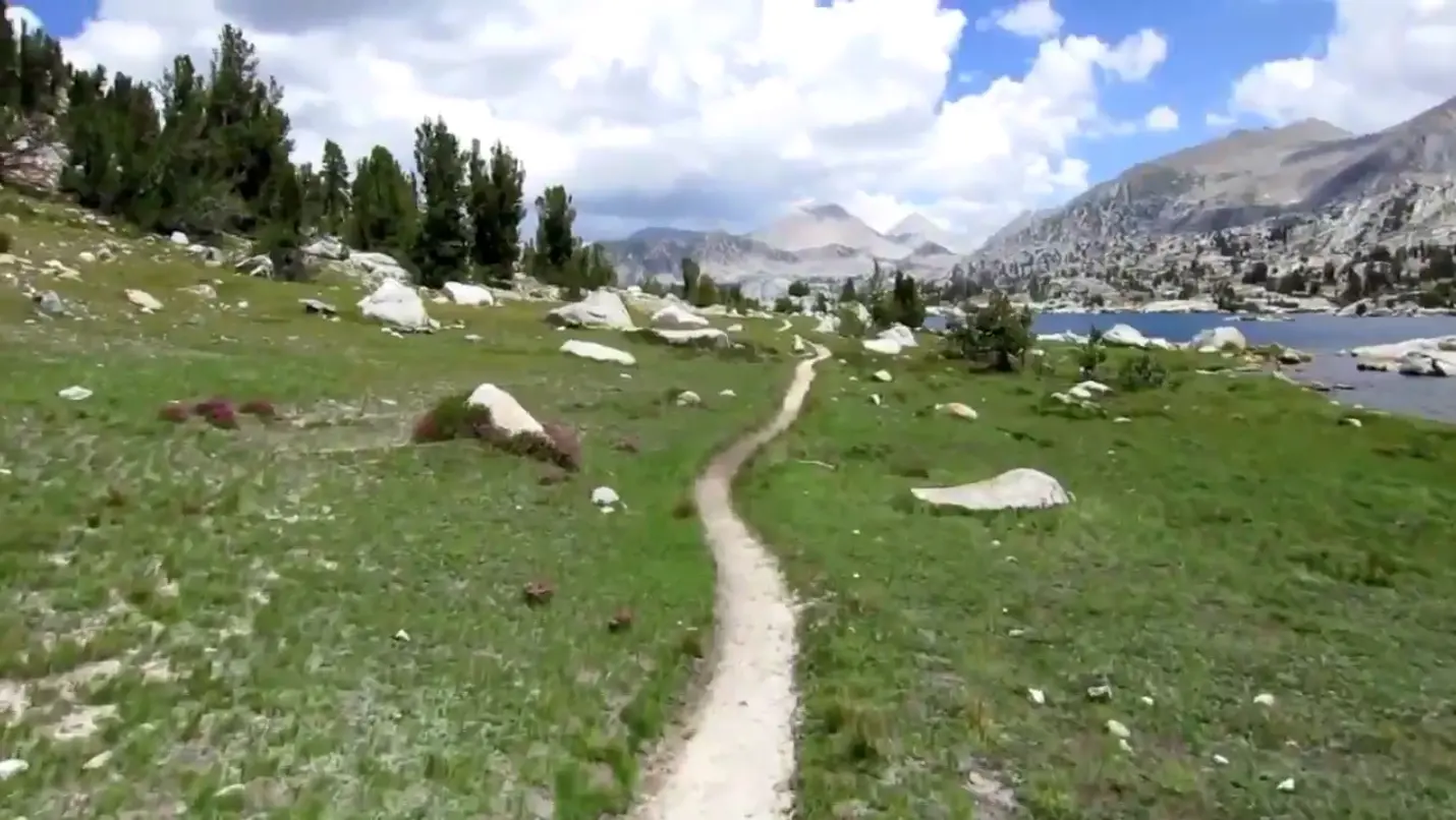 A narrow dirt trail winds through a green meadow dotted with rocks, inviting adventurers on a thru-hike towards distant mountains under a partly cloudy sky. Trees line the path, and a blue lake is visible on the right, perfect for capturing stunning PCT video memories.