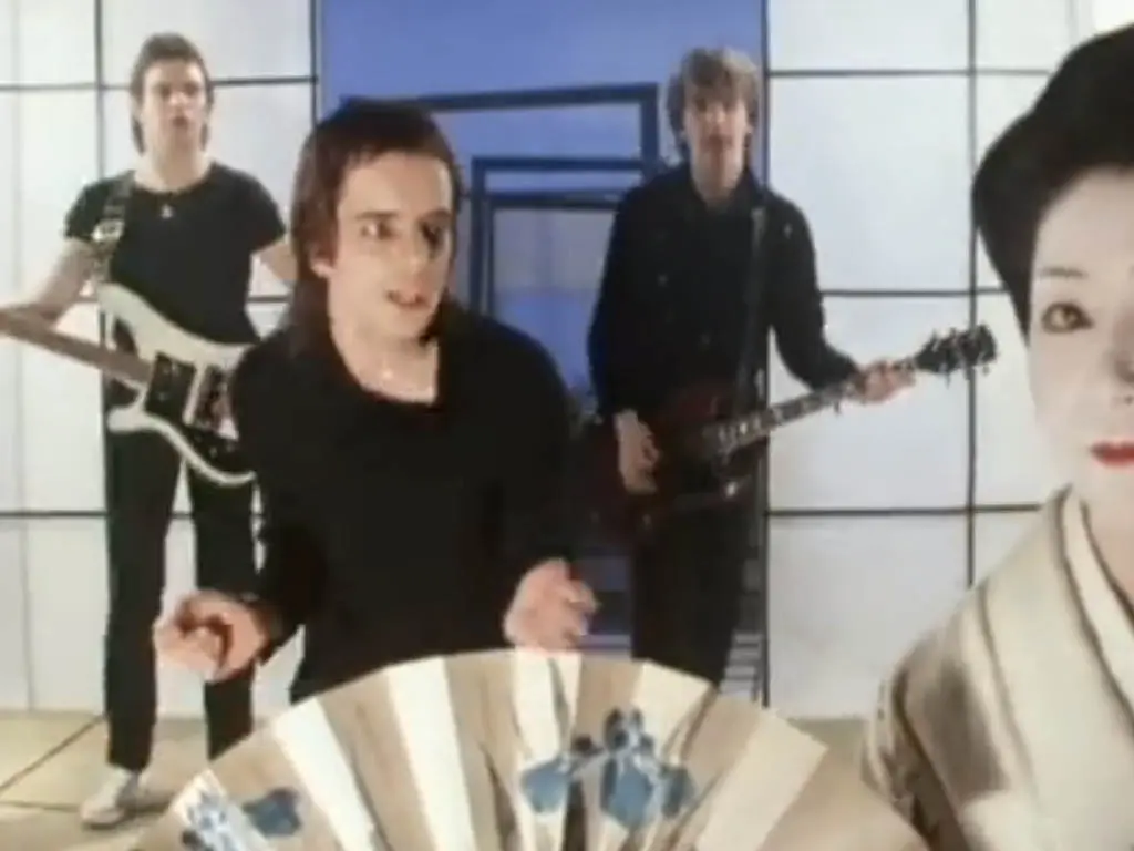 Four musicians perform indoors, evoking a vibe reminiscent of Turning Japanese. A person in traditional attire, holding a fan, stands prominently in the foreground on the right. The background is adorned with a geometric pattern of squares and rectangles.