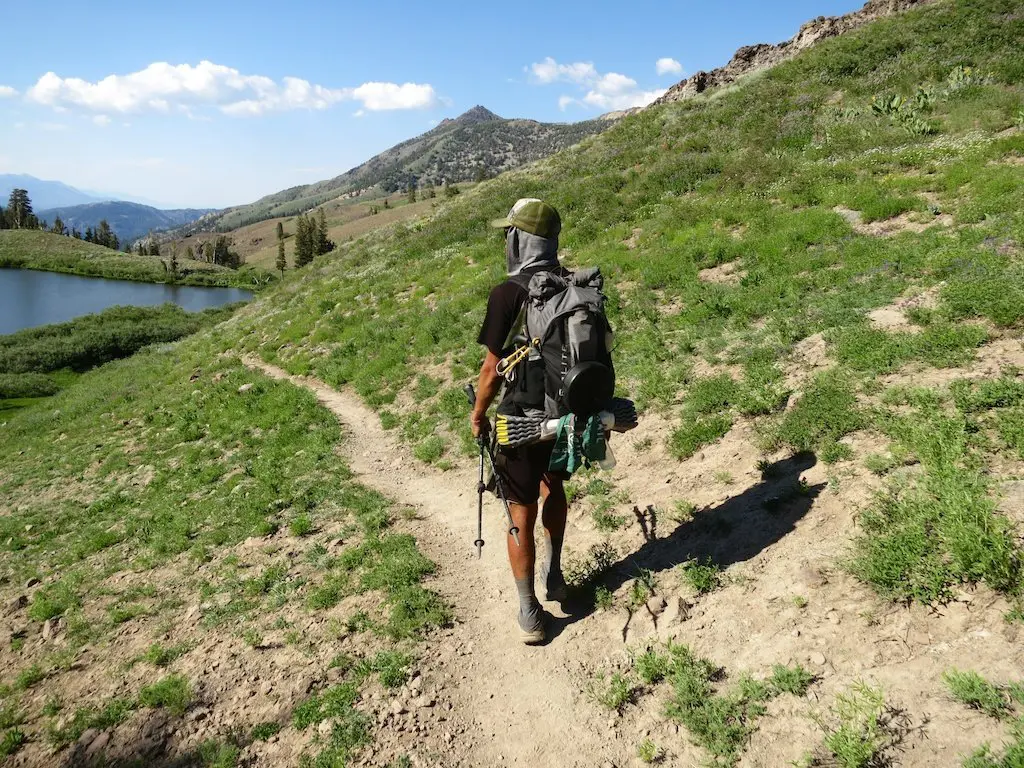 A hiker wearing a hat and mesh face covering strides along a dirt trail by a grassy hill, carrying a large backpack packed from their PCT gear list. A stunning lake and towering mountains stand in the background under a blue sky dotted with scattered clouds.
