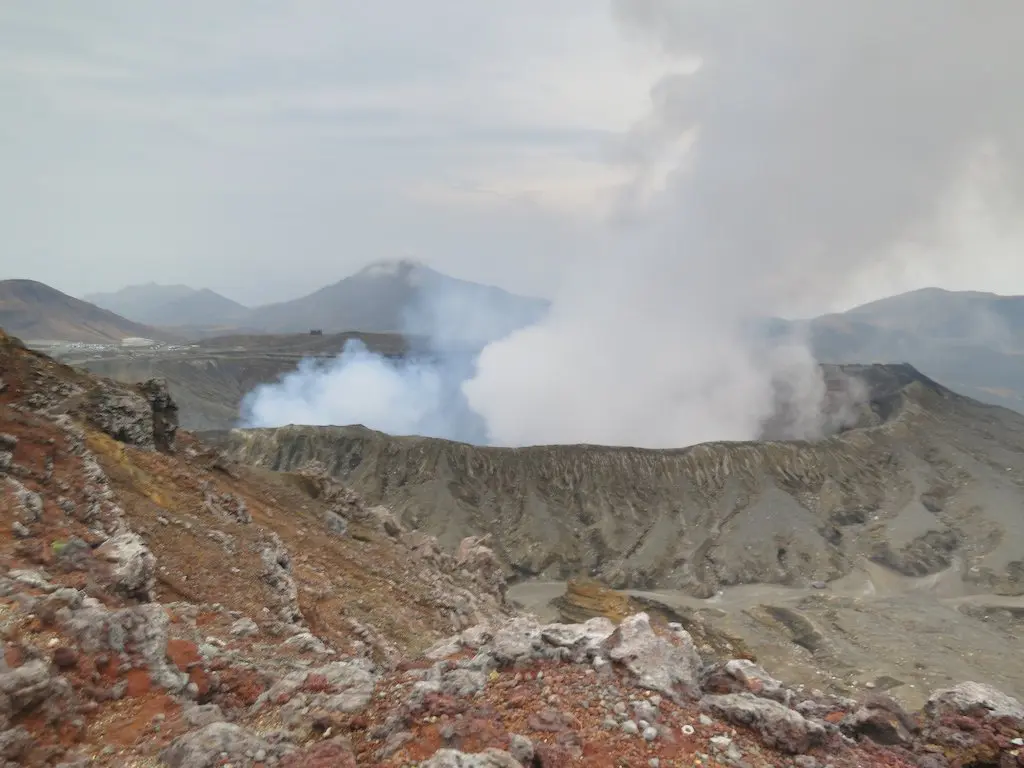 The Aso landscape boasts a volcanic crater emitting thick white smoke. In the foreground, reddish-brown rocky terrain contrasts with hazy mountain peaks that loom under a cloudy sky, painting a scene of raw and rugged beauty.