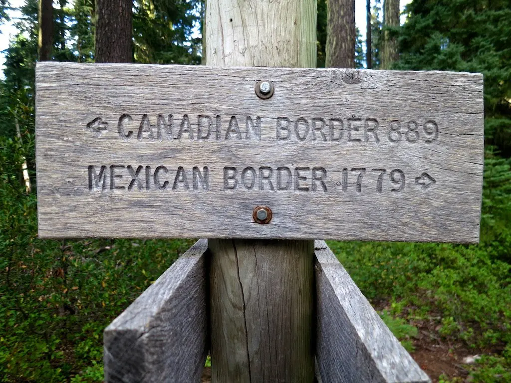 A weathered wooden signpost in the forest displays arrows for the Canadian Border 889 and the Mexican Border 1779. This Pacific Crest Trail marker, mounted on a sturdy wooden pole, is enveloped by greenery and towering trees.