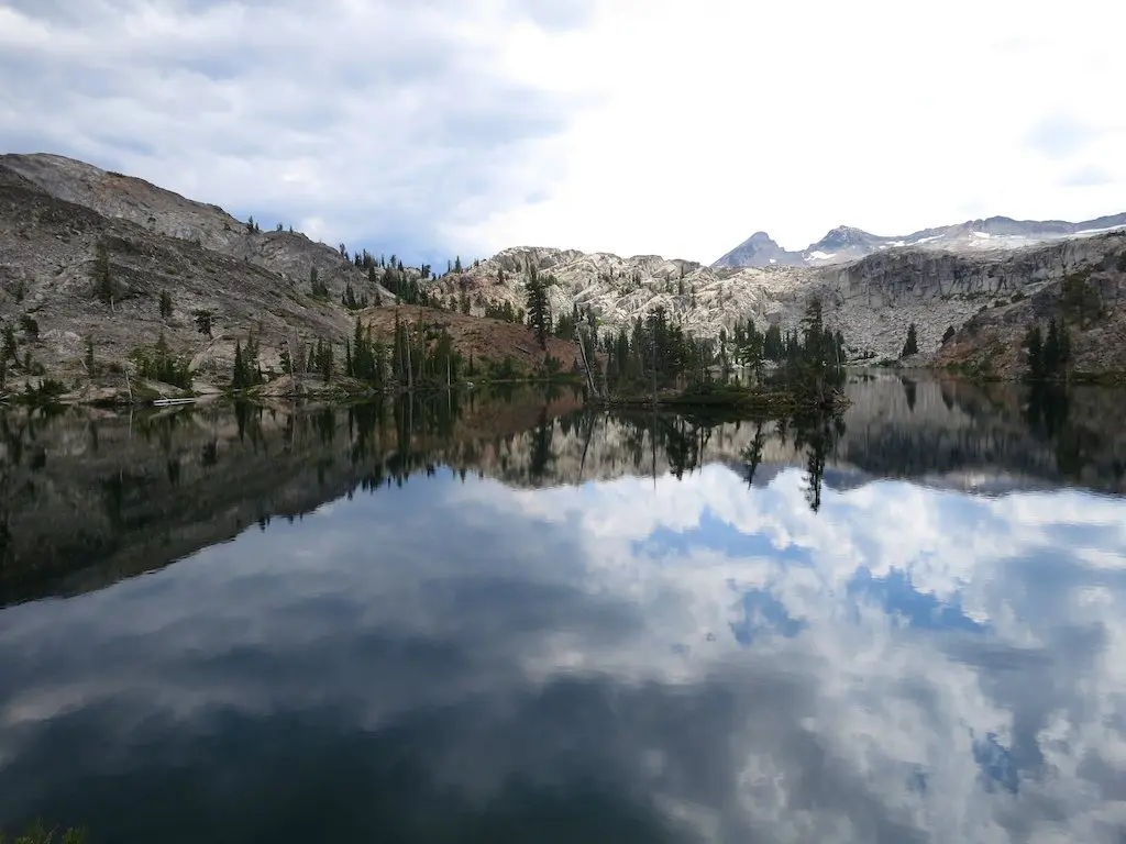 A tranquil NorCal lake mirrors a cloudy sky and the rugged mountains adorned with patches of pine trees. The serene landscape is painted in muted earth tones, with distant peaks standing under a bright, cloud-dappled sky.