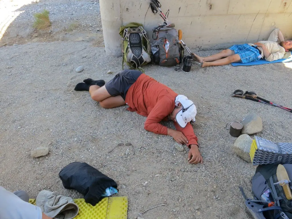 A person in a red shirt is sleeping on the ground under a bridge, using a white cap for shade—classic signs of a Pacific Crest Trail adventure. Camping gear, backpacks, and trekking poles are scattered nearby. Another trail enthusiast in blue shorts rests in the background.
