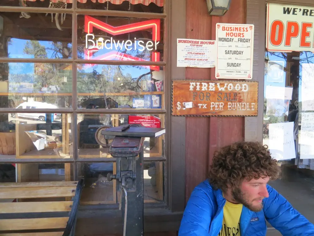 A man with curly hair in a blue jacket sits next to an old scale outside a rustic store. A Budweiser neon sign and various business signs adorn the window, listing firewood for sale and business hours. The scene feels timeless, as if unchanged despite any final pct stats or modern trends.