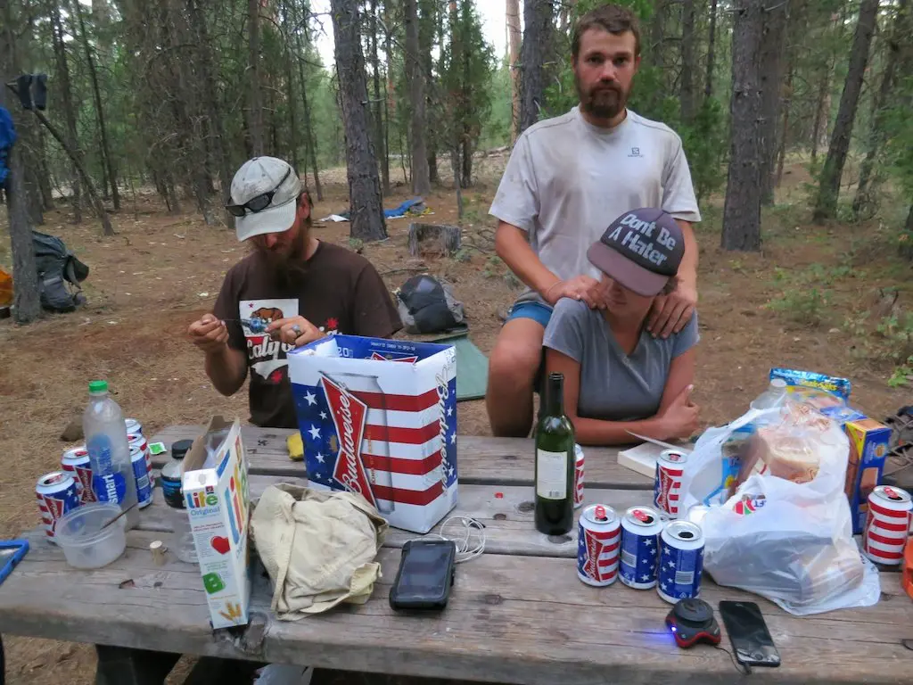 Three people at a picnic table in a forest, surrounded by food and drinks. One person is sitting with hands on anothers shoulders, who wears a hat like theyve been plucked from the pages of a Pacific Crest Trail glossary. Another is eating with a spoon amidst empty cans, a wine bottle, and a card game.