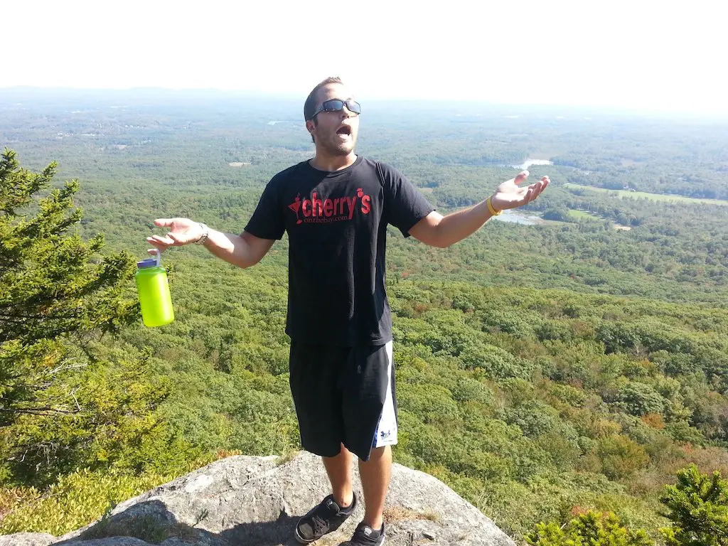 A man stands on a rocky ledge overlooking a vast green landscape, training for the Pacific Crest Trail. He is wearing sunglasses, a black T-shirt, and shorts, holding a yellow water bottle. With arms outstretched, he revels in the bright and clear sky above.