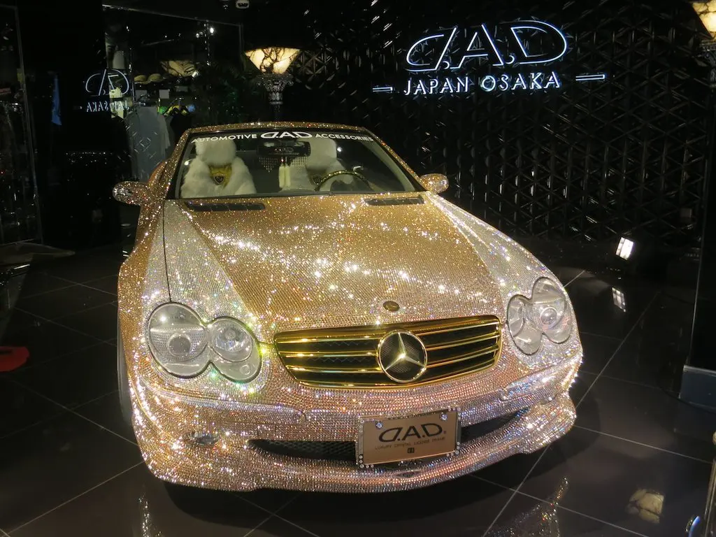 A luxury car adorned with sparkling crystals is on display in a showroom, exuding elegance under dim lighting. The car features a DAD logo on the front license plate and windshield, while a DAD Japan Osaka sign proudly hangs behind, celebrating its Japanese craftsmanship.