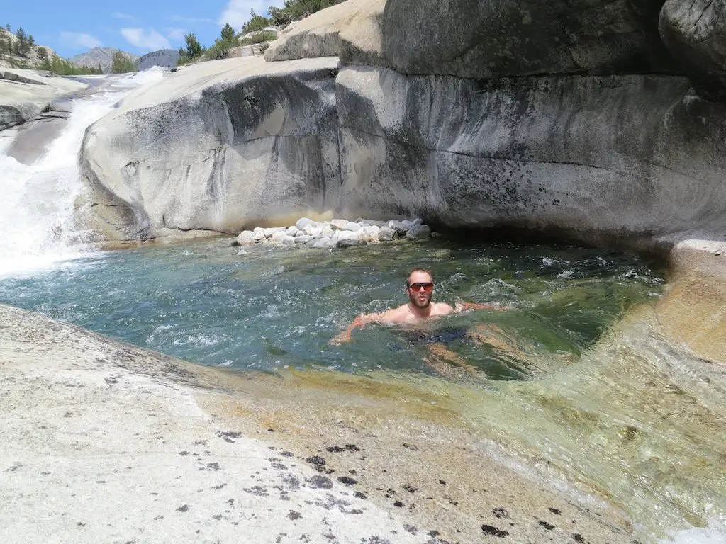 A person swimming in a natural rock pool with clear water, nestled along the scenic Pacific Crest Trail. The pool is surrounded by smooth rocks, and a small waterfall flows into it. Trees and mountains stand majestically in the background under a clear sky, offering a serene hiking paradise.