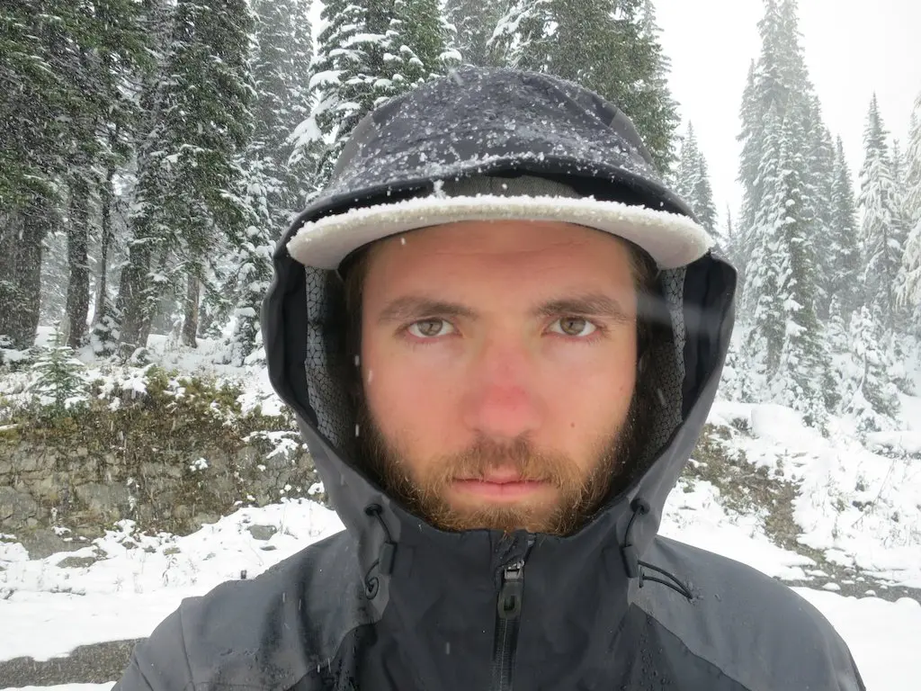 In a snowy landscape, a bearded thru-hiker stands bundled in a hooded jacket and cap, his face slightly red from the cold. Evergreen trees dusted with fresh snow form the backdrop, as if capturing a scene from an adventurous video journey.