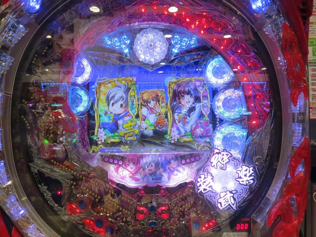 The pachinko machine dazzles with its bright LED lights, featuring vibrant images of three anime-style figures. Animated characters and decorative elements dance around a circular frame, where illuminated numbers and symbols add to the colorful spectacle.