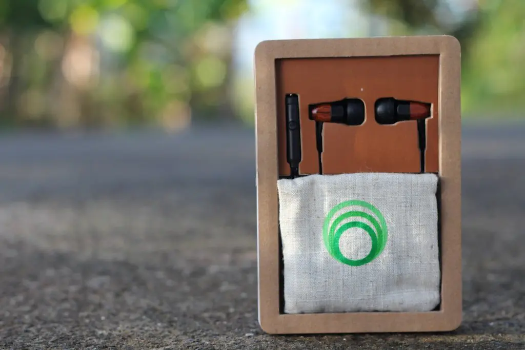 A pair of black and brown Thinksound TS02 earphones displayed in eco-friendly packaging, set against a blurred outdoor background. The package includes a small fabric pouch with a green circular logo on it.