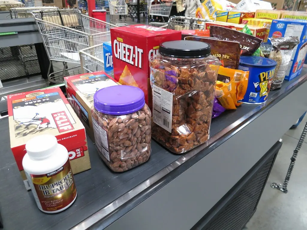 A grocery store checkout counter displays snack boxes, jars of nuts, ibuprofen, and a bag of Planters peanuts, sparking thoughts of the cost to hike PCT. Shopping carts and a barcode scanner linger in the background.
