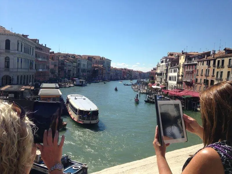 A vibrant canal in Venice, Italy, bustling with boats and gondolas, showcases why we often cant have nice things. Historic buildings with colorful façades line the waterway as two tourists capture the scene on tablets under a clear blue sky.