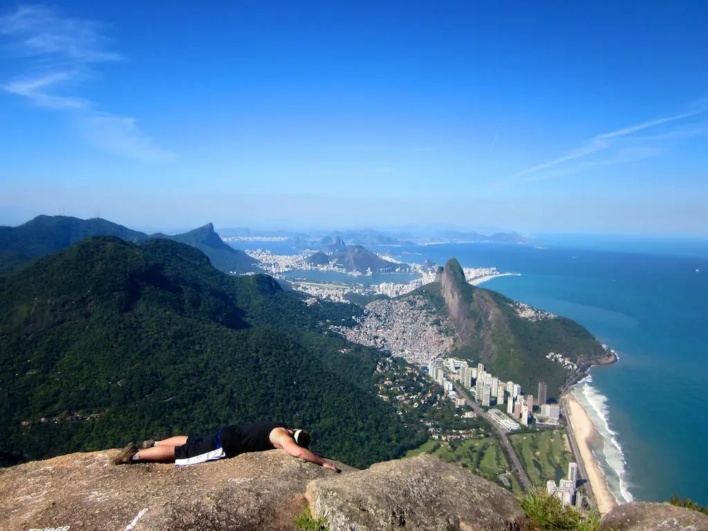 Someone lying on a rocky ledge overlooks a breathtaking landscape with lush mountains and a city by the ocean. Despite muttering I hate hiking, theyre captivated by the clear blue sky and sweeping view of the coastline with its sandy beaches.