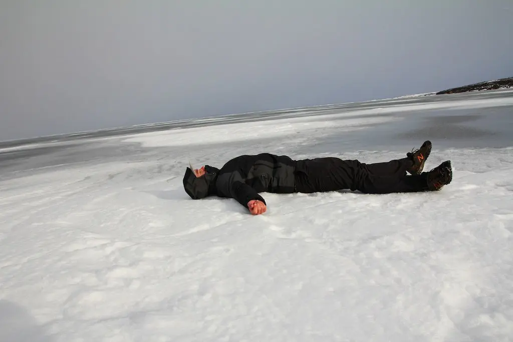 A person in dark winter clothing, perhaps from the Bluffworks Original collection, lies on their back on a snowy, icy landscape. The overcast sky looms above while the horizon stretches thin in the distance, painting a scene of cold desolation.