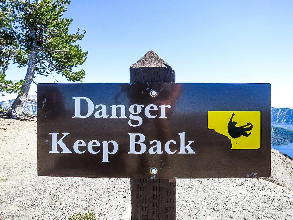 A sign with Danger Keep Back in white letters on a dark background depicts a silhouette of a person falling. Positioned near the cliff edge of the Pacific Crest Trail, it underscores trail safety amidst a stunning backdrop of a tree, lake, and distant mountains.